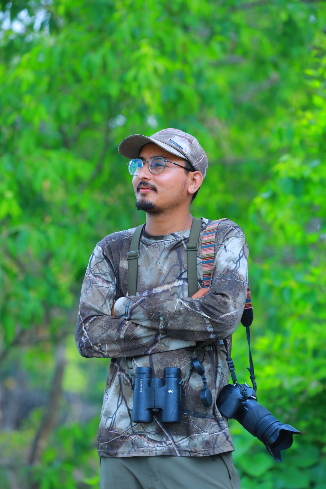 Krishna Thapa in camouflage field clothes with binoculars and a camera, arms crossed, in green forest