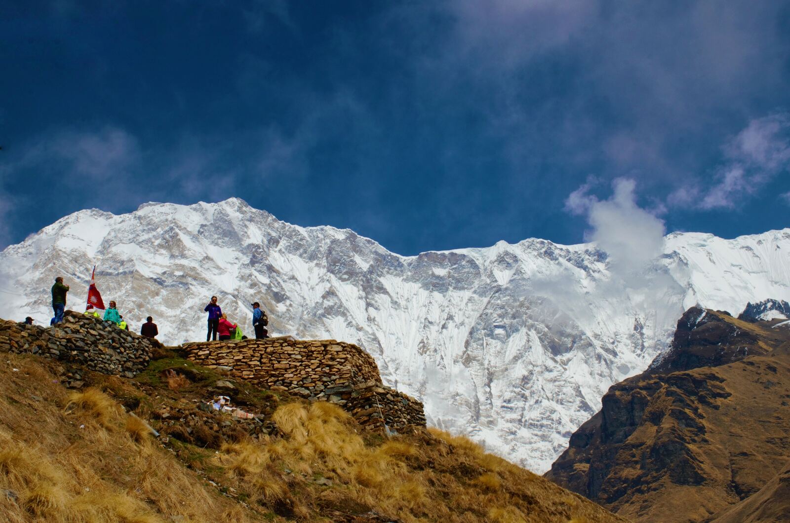 Trekkers on a rocky ledge below snow-capped Himalayan peaks under a blue sky